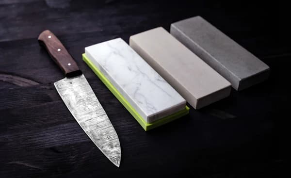 Damascus chef knife beside a three-stone whetstone progression on dark wood — 1000, 3000, 6000 grit